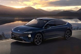A dark blue NIO ET9 electric car driving by a lake at sunset with mountains in the background.