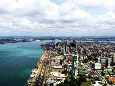 Aerial view of a coastal city with tall buildings, a busy road along the waterfront, and a large body of water under a partly cloudy sky.