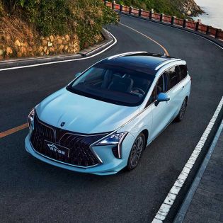 A light blue MPV drives on a coastal winding road, with lush greenery and the sea in the background, highlighting its sleek design and scenic appeal.