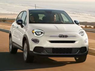 A white Fiat compact SUV drives along a coastal road, with sandy beaches and rolling hills in the background, highlighting its stylish design and scenic driving experience.