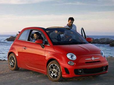 A red Fiat 500 parked by the seaside with two people interacting around it.