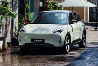 A light green Geely Xingyuan electric car parked on a wet brick road with plants and umbrellas in the background.