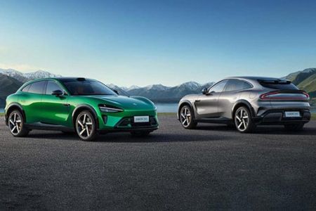 Two Xiaomi electric cars, one green and one silver, parked on a paved area with a scenic backdrop of mountains and a lake.
