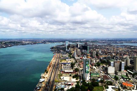 Aerial view of a coastal city with tall buildings, a busy road along the waterfront, and a large body of water under a partly cloudy sky.