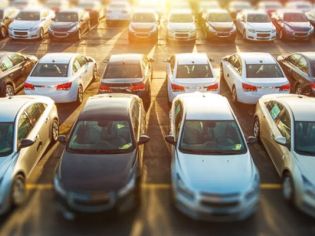 Sunlit parking lot filled with rows of parked cars, featuring a mix of white, black, and silver vehicles.