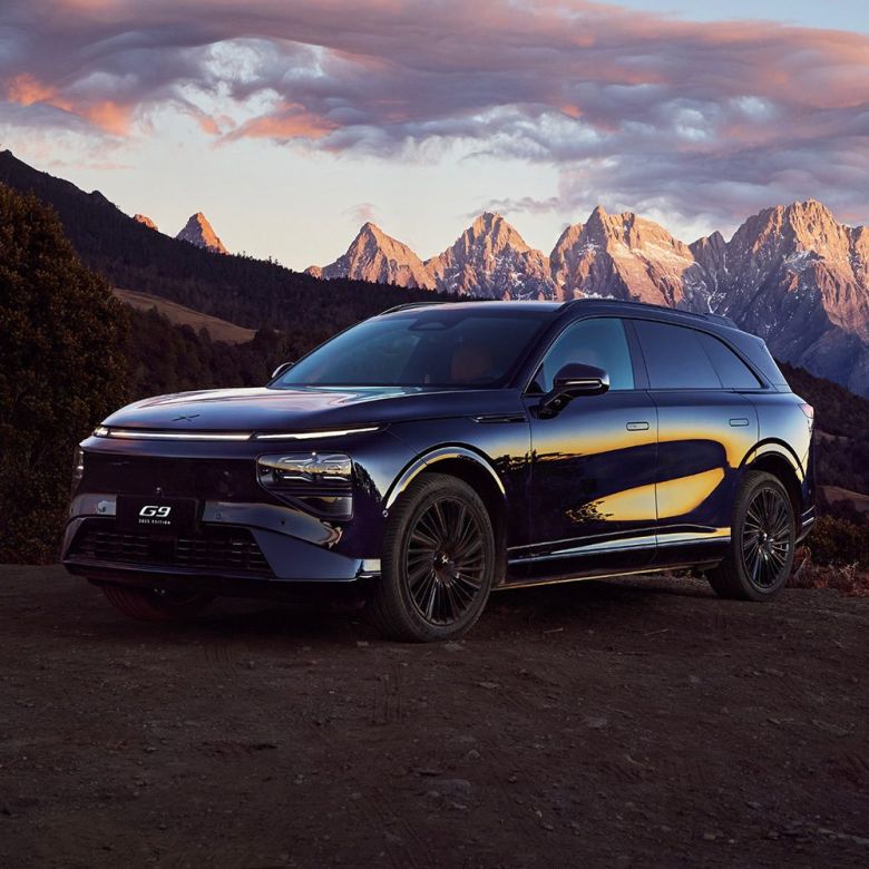 A dark - colored Xpeng G9 SUV parked with a stunning mountain range and a colorful sunset sky in the background.