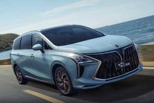 A light blue MPV drives along a coastal road, with the sea and clear sky in the background, showcasing its sleek design and dynamic appeal.