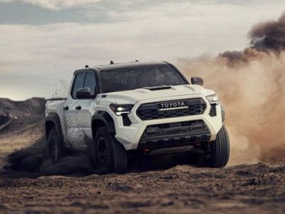 A white Toyota pickup truck drives on a dusty desert terrain, kicking up a cloud of dirt, with rugged hills and a partly cloudy sky in the background.