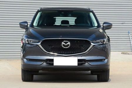 Front view of a dark gray Mazda SUV, parked in front of a corrugated metal wall, featuring its grille, headlights, and sleek front design with a blank license plate.