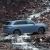 A light blue SUV driving through a stream in a rocky, mountainous area.