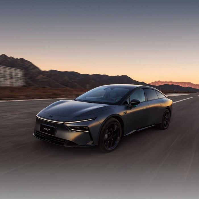 A dark gray Xpeng P7+ electric car driving on a road with mountains and a sunset sky in the background.