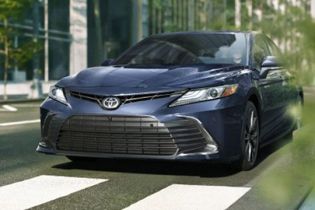 Front view of a dark blue Toyota Camry sedan driving on a city street, with modern buildings and greenery in the background, highlighting its sleek urban design.