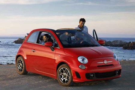 A red Fiat 500 parked by the seaside with two people interacting around it.