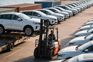 A forklift handling a car at a port with rows of white cars and ships in the background.