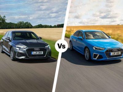 Two Audi sedans (black vs bright blue) on a rural road: side-by-side "vs" comparison. Both modern, sleek models (likely A3/A4 variants) driving through countryside with fields/trees in background. License plates: LJV 360 (black), JJF 500 (blue).