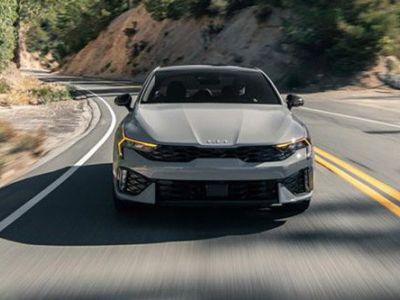 A gray Kia sedan driving on a winding mountain road, with lush greenery and rocky terrain in the background, highlighting its dynamic design and performance.