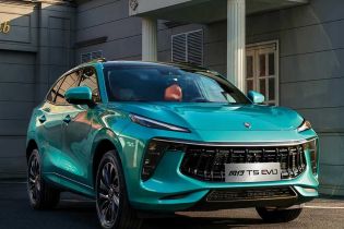 A turquoise Fengxing T5 EVO SUV parked on asphalt near a gray building with autumn trees in the background, showcasing its sleek and modern front design.
