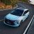 A light blue MPV drives on a coastal winding road, with lush greenery and the sea in the background, highlighting its sleek design and scenic appeal.