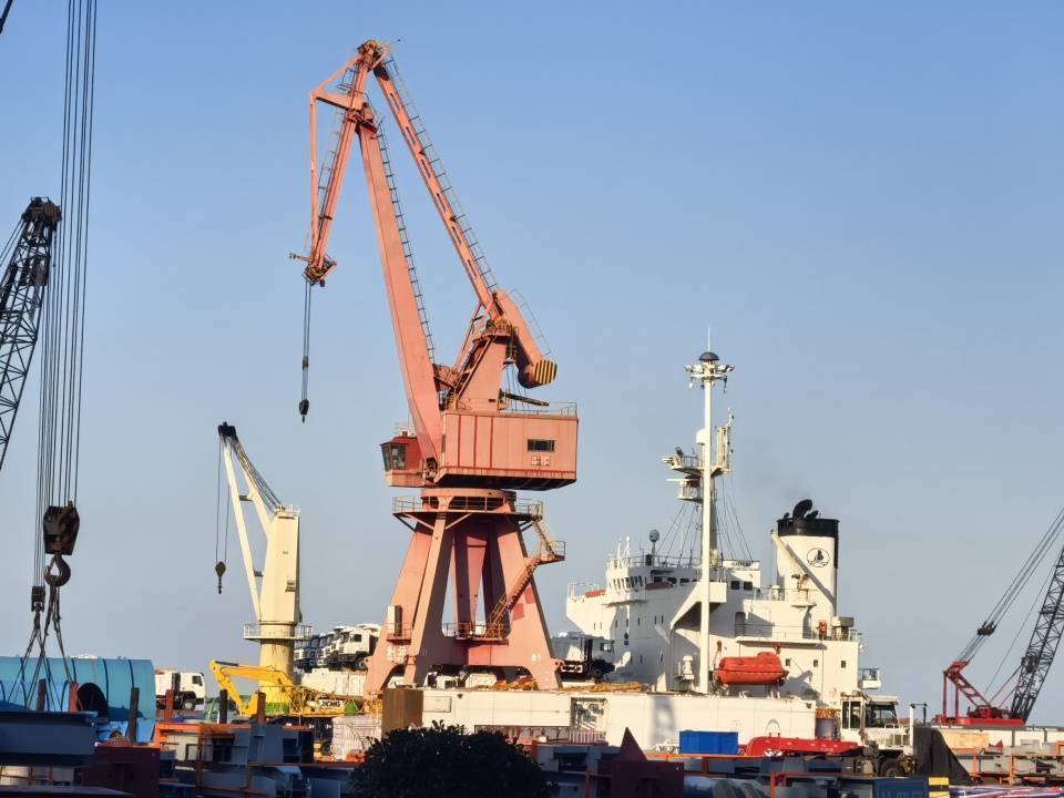 A large red crane and a ship at a busy port under a clear blue sky.