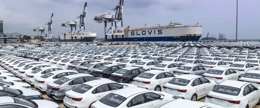 A large number of cars parked at a port with a "GLOVIS" ship and cranes in the background.
