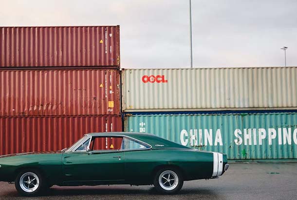 A green vintage car parked in front of stacked shipping containers with "OOCL" and "CHINA SHIPPING" logos.