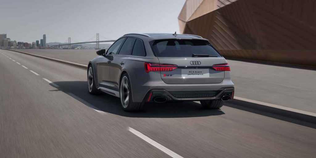 A silver Audi RS 6 Avant performance wagon driving on a road with an urban background and a bridge.