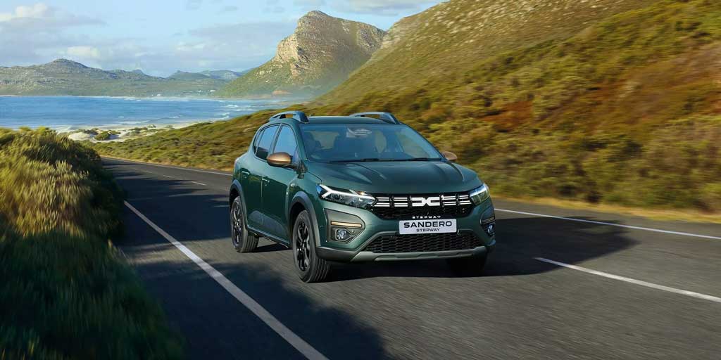 A green Dacia Sandero Stepway driving on a road by the seaside with a scenic background.