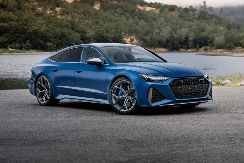 A blue Audi car parked by a lake with a forested hillside in the background.