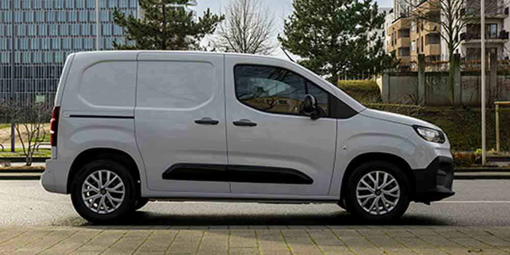 Side view of a silver van parked on a street in an urban area with modern buildings and trees in the background.
