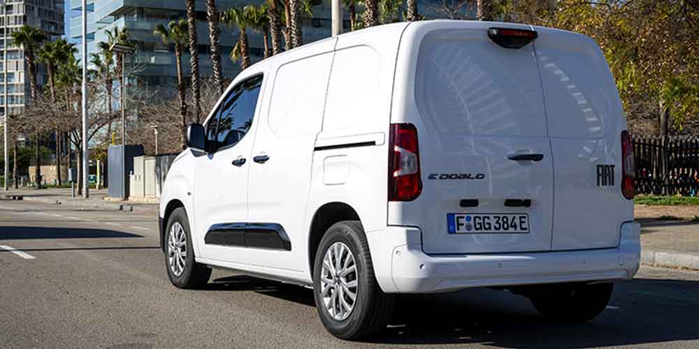 Rear view of a white Fiat E - Doblò van with license plate F GG 384E, parked on a street in an urban area with palm trees and modern buildings in the background.