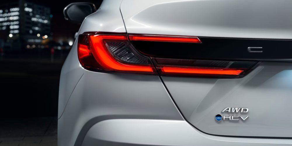 Close-up of the rear section of a white car, focusing on its red tail light and "AWD HEV" badge, set against a nighttime urban backdrop, showcasing its hybrid and all-wheel-drive features.