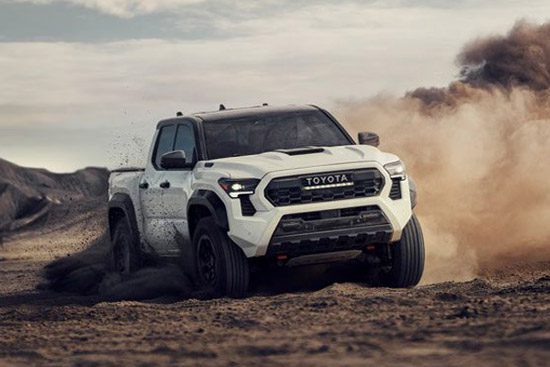 A white Toyota pickup truck drives on a dusty desert terrain, kicking up a cloud of dirt, with rugged hills and a partly cloudy sky in the background.