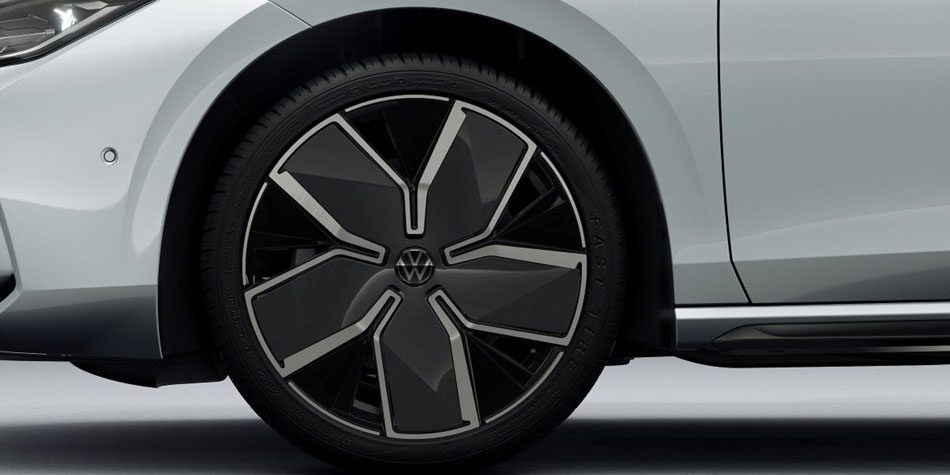 Close-up of a light gray Volkswagen car’s wheel: Features a black-and-silver multi-spoke alloy rim with the VW logo at the center. Shows the wheel’s modern, geometric design against the car’s body.