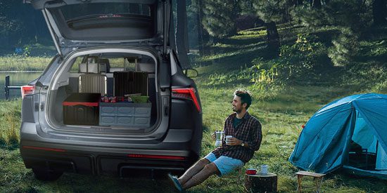 A man relaxes by a blue tent; a SUV’s open trunk holds camping gear, set in a grassy, wooded area.
